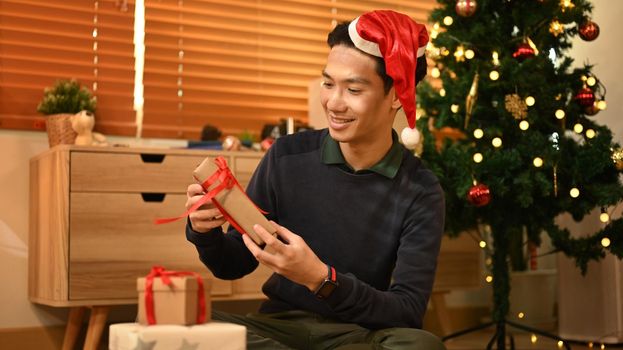 Smiling man wearing Santa hat sitting on floor in cozy living room and preparing Christmas gifts.