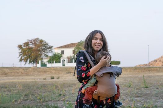 Young mother with her toddler kid boy on back in ergonomic baby carrier in autumn nature. Active mother concept
