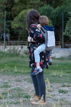 Young mother with her toddler kid boy on back in ergonomic baby carrier in autumn nature. Active mother concept