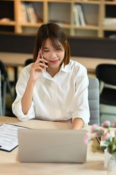 Pretty young woman entrepreneur having phone conversation with her partner and using laptop on wooden table.