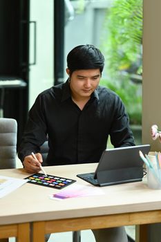 Young man graphic designer working with digital tablet and color palette, sketching and planning project on wooden office desk.