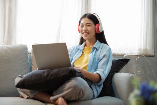 Young asian woman smiling listening to music using laptop surfing the internet while sitting on couch at home. Online connection and online learning concept.