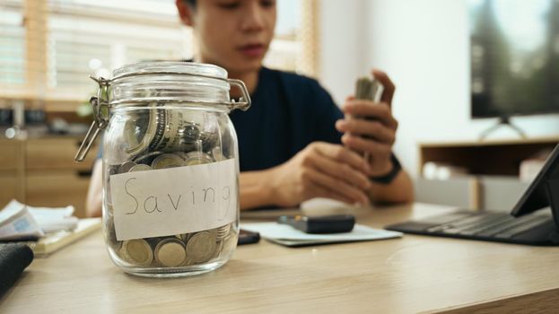 A glass jar with dollar bills and coins on wooden table, man using laptop in background. Saving money and investing concept.