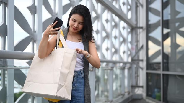 Trendy asian woman holding smart phone and shopping bags standing on terrace of modern building. Lifestyle concept.