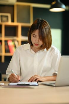 Concentrated working woman using laptop and signing business document on wooden table.