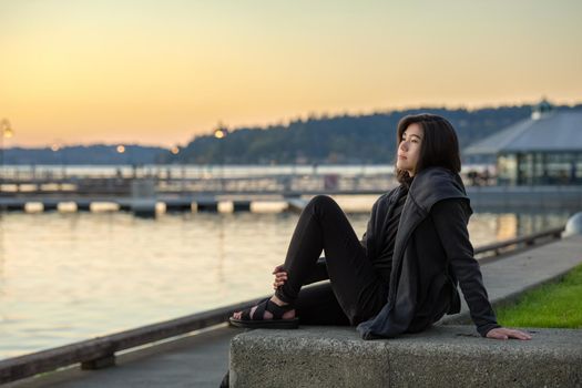 Smiling teen girl or young adult female in gray jacket sitting outdoors by lake enjoying colorful sunset on cool evening