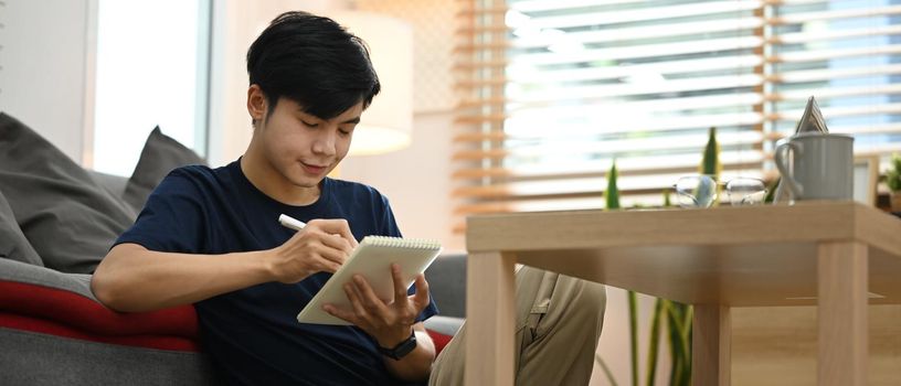 Handsome man freelancer sitting front of laptop in cozy living room and making notes on notebook.