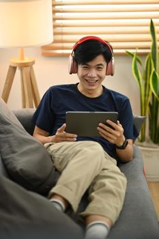 Happy man wearing wireless headphone and browsing internet on digital tablet while resting in cozy living room.