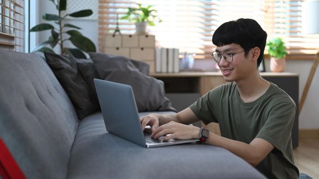 Smiling asian man sitting on floor in cozy living room and sending email or working online from home with laptop.