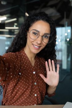 Vertical shot, young beautiful Hispanic business woman with curly hair talking on video call, waving at smartphone camera, using remote communication app