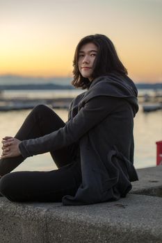Smiling teen girl or young adult female in gray jacket sitting outdoors by lake enjoying colorful sunset on cool evening
