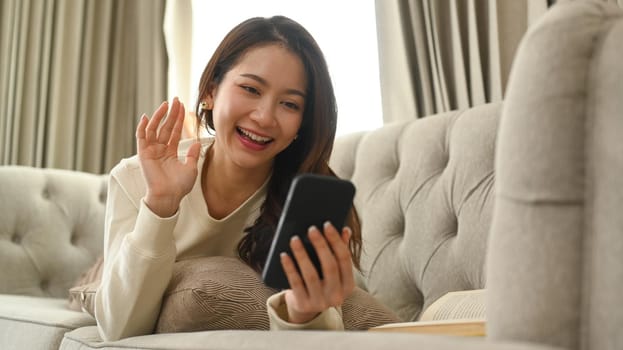 Cheerful young woman waving hands and making video call via smart phone while resting on couch at home.