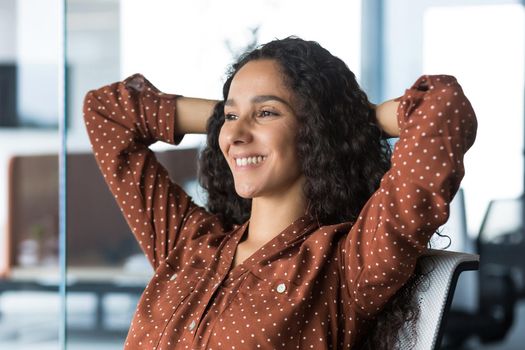 Close-up photo of a young beautiful business woman entrepreneur resting in the office looking out the window with her hands behind her head, businesswoman working on a laptop smiling and dreaming.