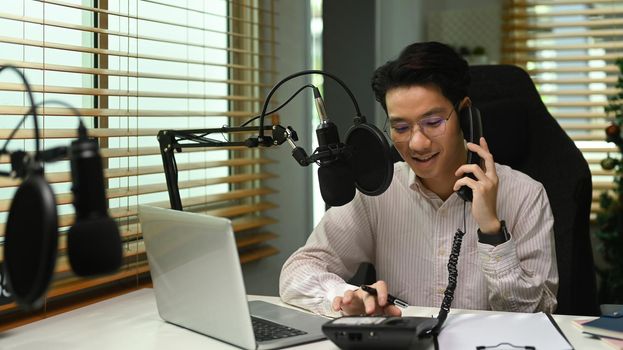 Asian man radio host talking on telephone and using condenser microphone recording voice conversation at home studio.