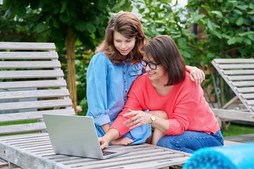 Middle aged mom and preteen daughter looking at laptop together, outdoor on armchairs in backyard. Parent and child hugging using laptop to study leisure shopping