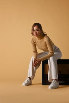 Attractive fashionable woman wearing trendy sweater sitting on a vintage radio, studio shot on beige background.