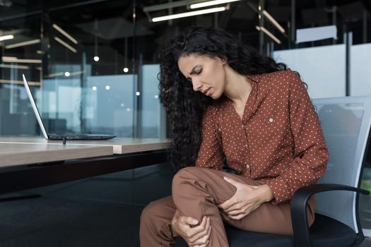 Young beautiful arab woman working in modern office with laptop, curly business woman has severe pain in leg muscles, varicose veins, massaging her leg