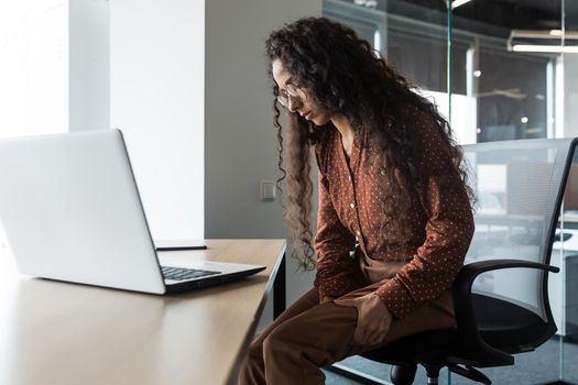 Young beautiful arab woman working in modern office with laptop, curly business woman has severe pain in leg muscles, varicose veins, massaging her leg