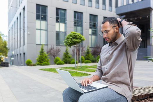 Serious and pensive man sitting on bench unhappy with work result, using laptop for remote work, african american businessman outside office building sitting on bench.