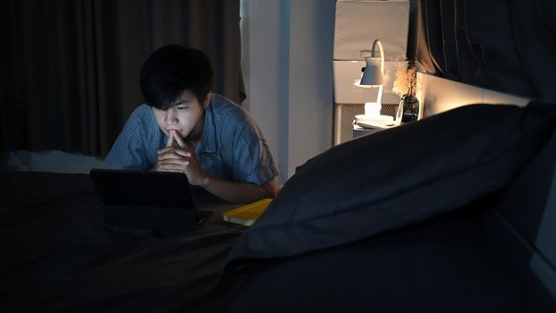 Young asian man lying on his bed and surfing inter.net with computer laptop.