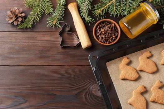 Ready-made Christmas cookies in the form of a rabbit on a baking sheet on a wooden table decorated with Christmas tree branches, with a jar of honey and cloves. Copy space