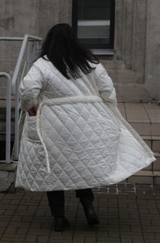 A plus-size woman with black hair in a white coat walks down the street back view.