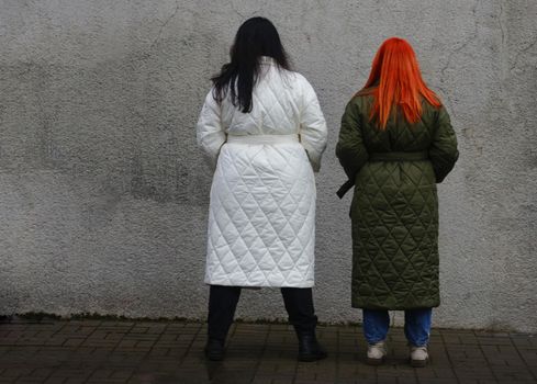 A brunette in a white coat and a brown-haired woman in a brown coat pose against a gray old concrete wall. view from the back.