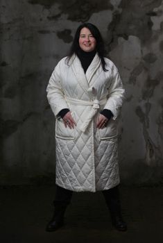 Portrait of a woman plus size model with dark hair in a white XXL winter coat and black trousers posing against the wall.