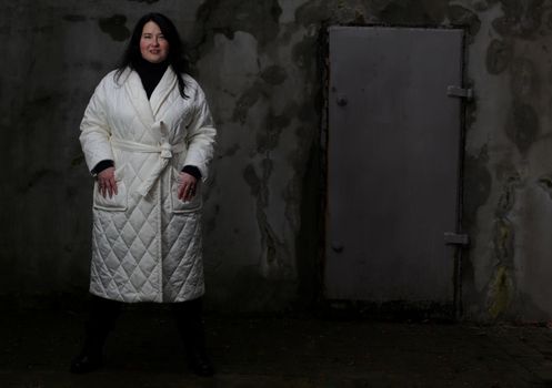 Portrait of a woman plus size model with dark hair in a white XXL winter coat and black trousers posing against the wall.