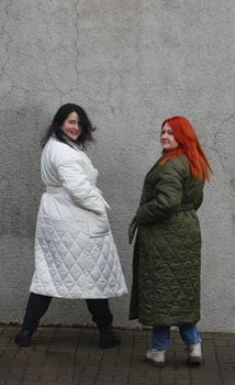 Two girls in plus-size clothes. Two girls in white and green coats in front of a wall with old gray stucco turn around and look at the camera.