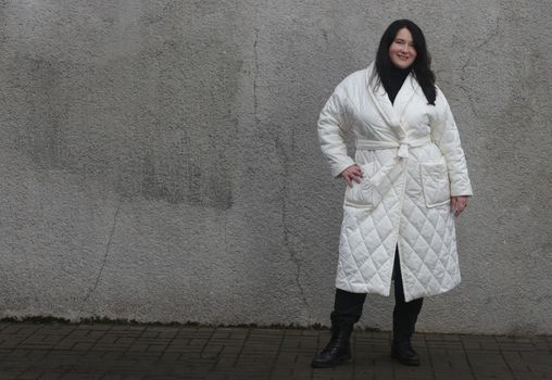 Portrait of a woman plus size model with dark hair in a white XXL winter coat and black trousers posing against wall.