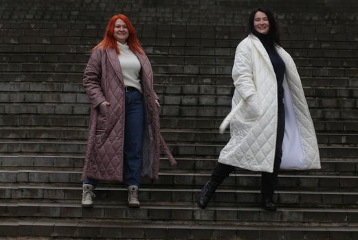 A woman with red hair in a brown coat and a woman with black hair in a white coat. Two women plus size in unbuttoned raincoats.