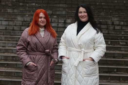 Two plus-size women posing in coats against a staircase backdrop...Brunette in white coat and brown-coat brown-haired woman look at each other in front of the camera. Two women in plus-size winter clothes.
