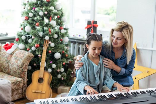 Merry Christmas and Happy Holidays. Cheerful mom and her cute daughter girl playing on a white piano against the background of a decorated Christmas tree. New Year.