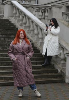 A brown-haired woman in a red coat and a brunette in a white coat lean on the large railing of the stairs and hold a smartphone.