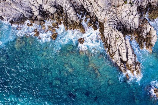 aerial top view of clear turquoise ocean with sea waves hitting and crashing on the cliffs of the seashore of the rocky coast with spray and foam, power of the nature concept