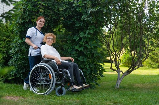 Caucasian female doctor walks with an elderly patient in a wheelchair in the park. Nurse accompanies an old woman on a walk outdoors