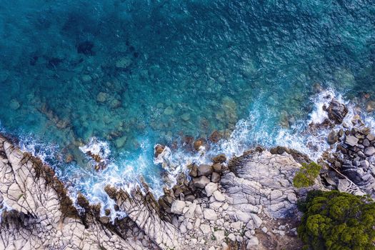 top view of clear blue ocean with sea waves hitting on the cliffs of the seashore of the rocky coast with spray and foam, power of the nature concept