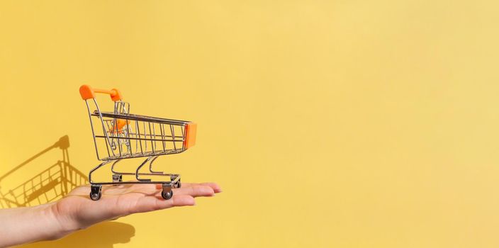 On a woman's hand is a small empty metal trolley from a supermarket. Banner. The object is on a yellow background. Horizontal photo. trading concept. Buyer. Shopping concept. Close-up. Copy space.