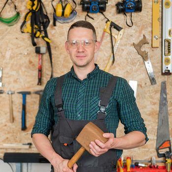 Portrait of a carpenter in goggles and overalls holding a wooden hammer in the workshop against the background of a wall with tools