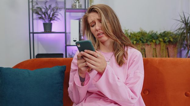 Portrait of caucasian adult girl use smartphone typing browsing, loses becoming surprised sudden lottery results, bad news, fortune loss, fail. Young woman sitting on orange couch at home living room