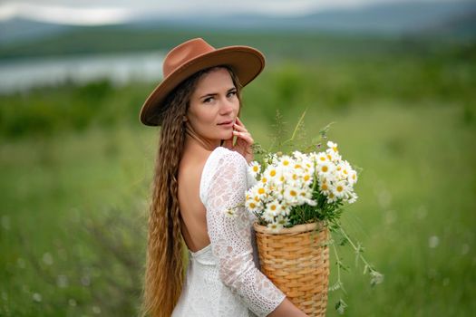 A middle-aged woman in a white dress and brown hat stands on a green field and holds a basket in her hands with a large bouquet of daisies. In the background there are mountains and a lake