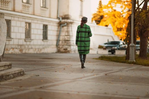 Outdoor fashion portrait of an elegant fashionable brunette woman, model in a stylish cap, green dress, posing at sunset in a European city in autumn