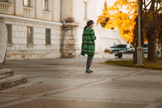 Outdoor fashion portrait of an elegant fashionable brunette woman, model in a stylish cap, green dress, posing at sunset in a European city in autumn