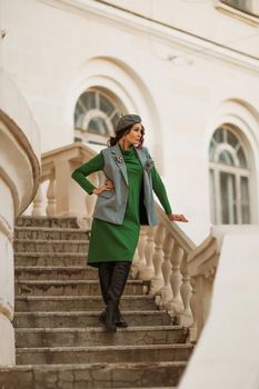 Outdoor fashion portrait of an elegant fashionable brunette model in a beret, green dress and a gray waistcoat posing at sunset in a european city in autumn