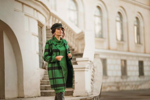 Outdoor fashion portrait of an elegant fashionable brunette model in a beret, green dress and a gray waistcoat posing at sunset in a european city in autumn