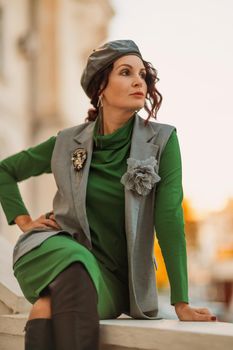 Outdoor fashion portrait of an elegant fashionable brunette model in a beret, green dress and a gray waistcoat posing at sunset in a european city in autumn