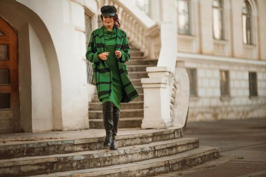 Outdoor fashion portrait of an elegant fashionable brunette woman, model in a stylish cap, green dress, posing at sunset in a European city in autumn