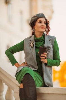 Outdoor fashion portrait of an elegant fashionable brunette model in a beret, green dress and a gray waistcoat posing at sunset in a european city in autumn