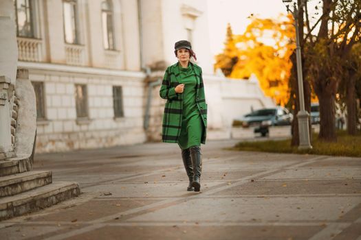 Outdoor fashion portrait of an elegant fashionable brunette woman, model in a stylish cap, green dress, posing at sunset in a European city in autumn